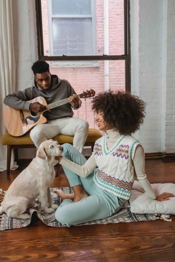 happy young ethnic woman stroking pet on floor near boyfriend playing guitar